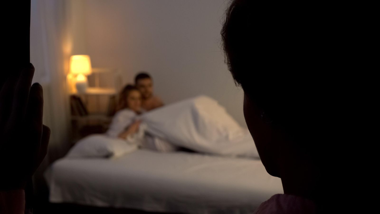 Una parella en el llit mentre una persona observa des de la porta