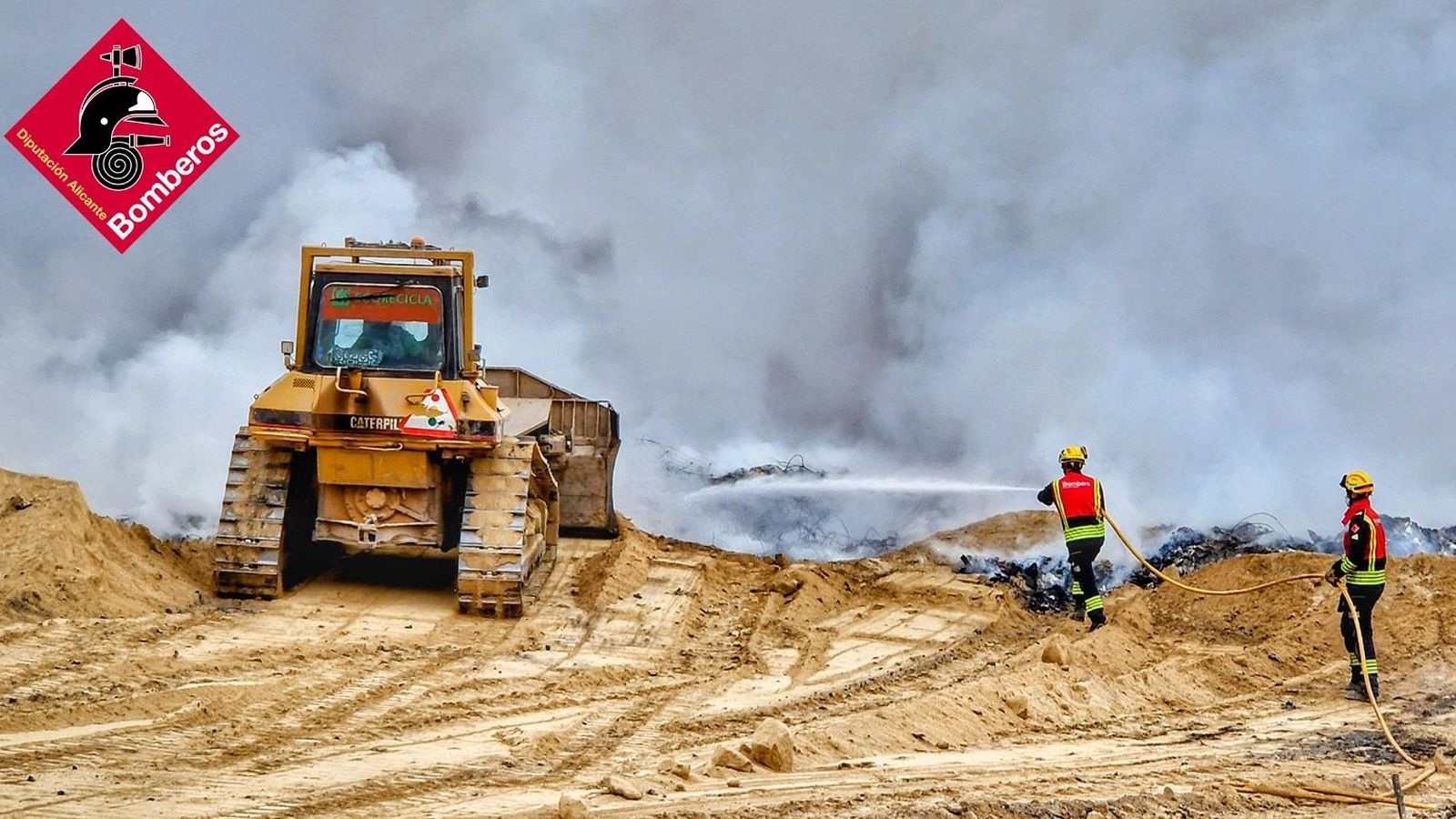 Un tractor i dos bombers intenten cobrir amb terra els muntons de residus cremats de la planta de reciclatge de Xixona