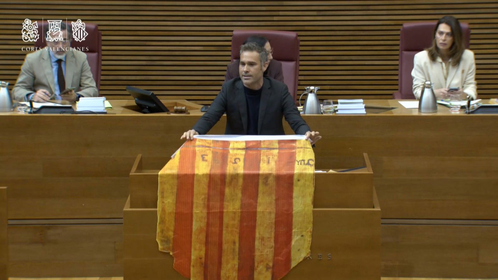 El síndic de Compromís Gerard Fullana exhibix un 'Penó de la Conquesta" de Jaume I a València en el ple de les Corts