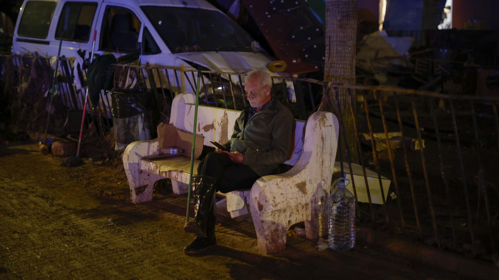 Un home consulta el seu telèfon en un banc d'un carrer a Paiporta, a l’Horta Sud, este divendres.