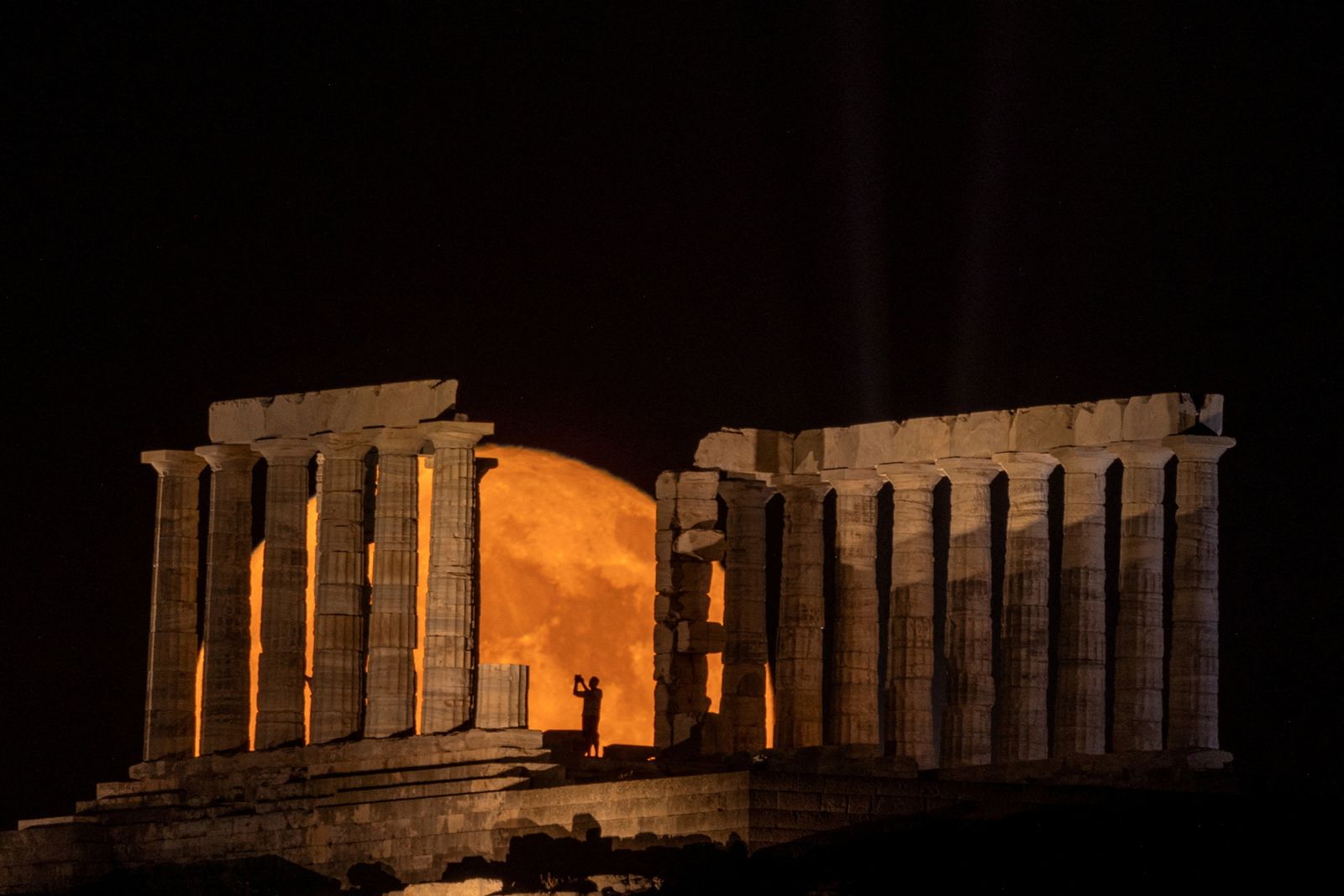 Una persona pren una fotografia mentre la lluna plena s'eleva darrere del temple de Posidó, prop d'Atenes, Grècia, 3 de juliol del 2023