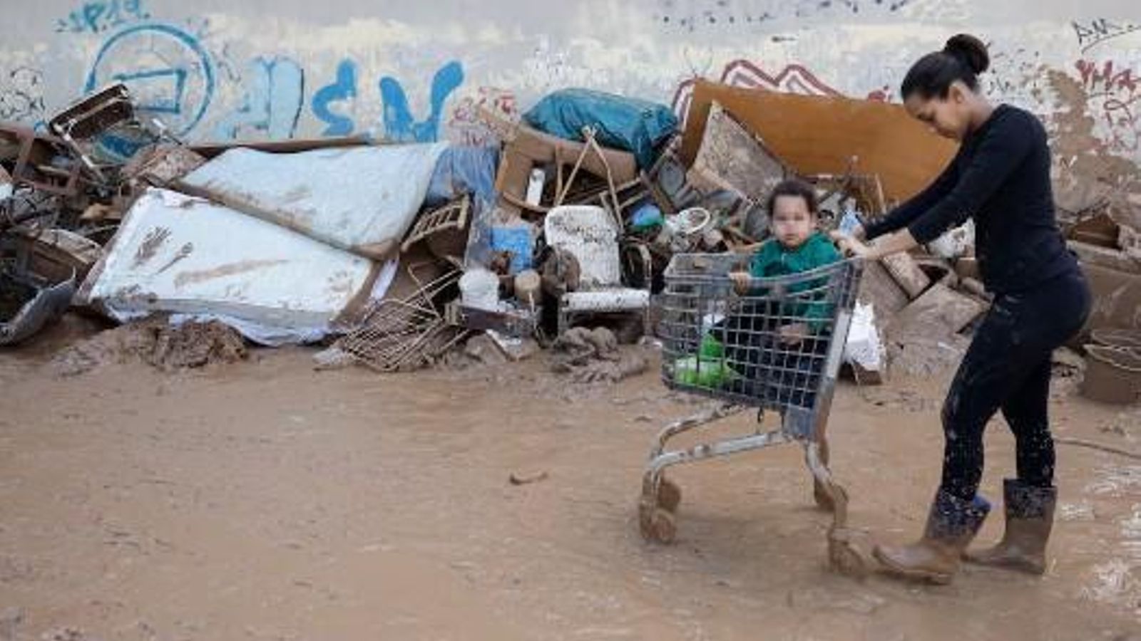 Una dona porta a un xiquet en un carro de la compra per un carrer ple de llot a Massanassa