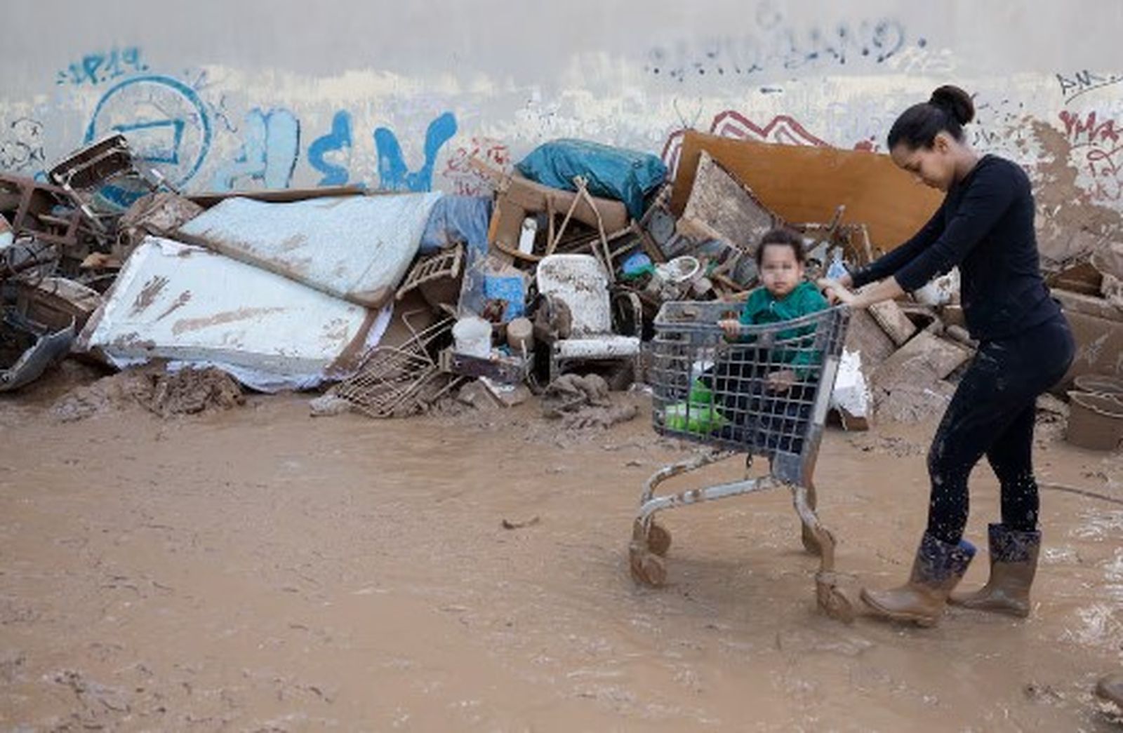 Una dona porta un xiquet en un carro de la compra per un carrer ple de fang a Massanassa