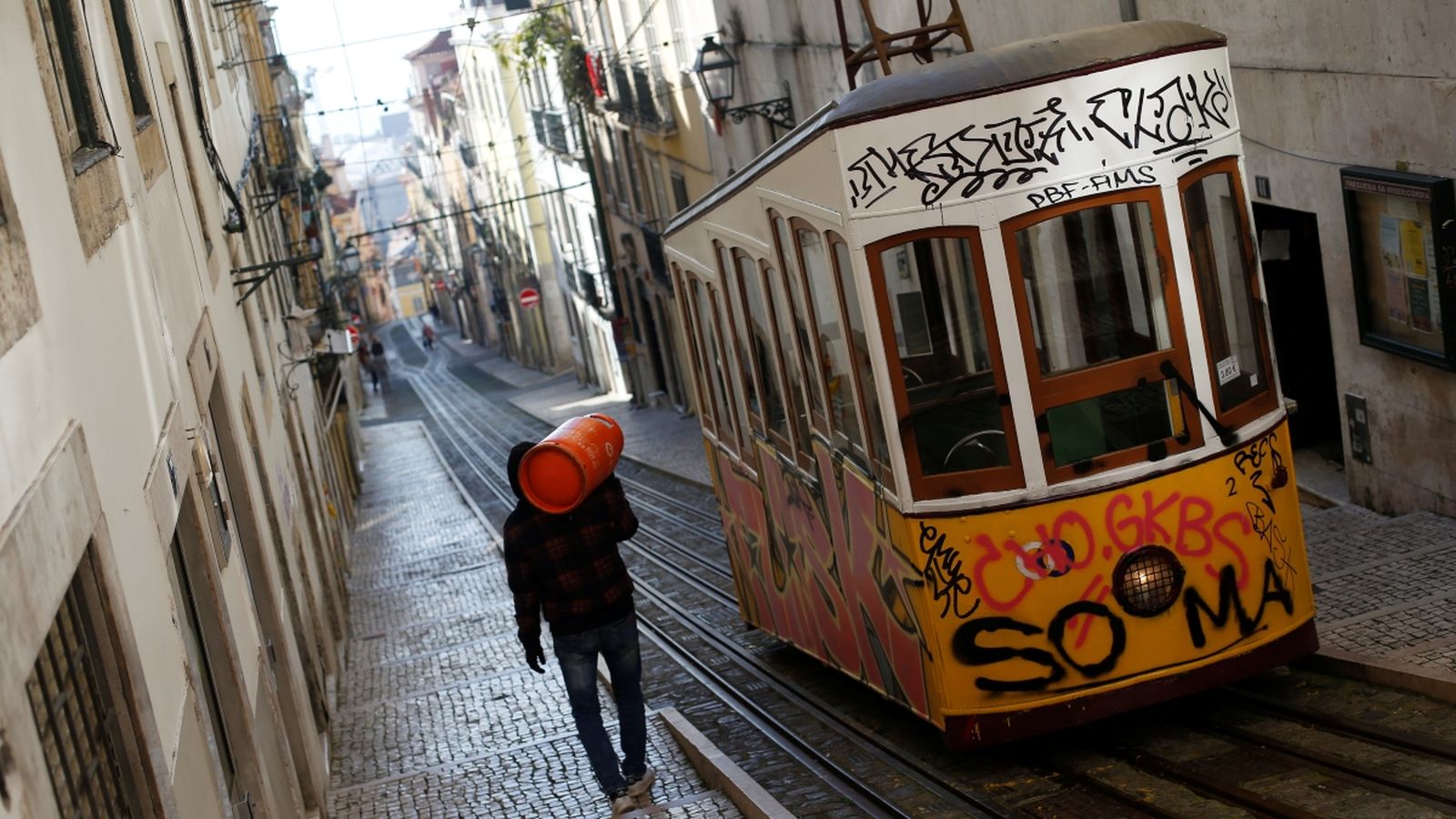 Un home carrega una bombona de butà pel carrer, mentre el tramvia passa pel seu costat