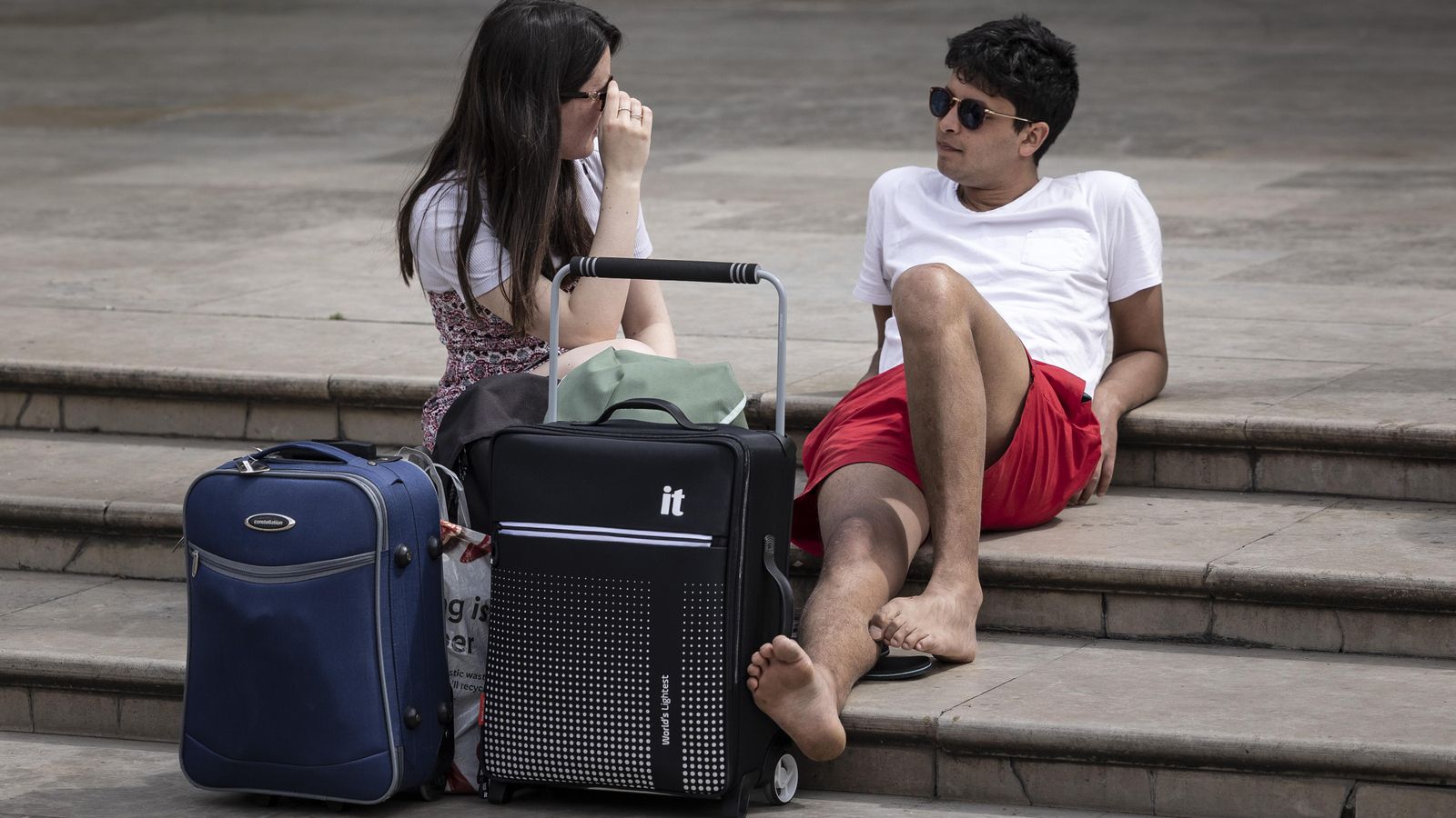 Dos turistes descansen amb les seues maletes a la plaça de la Mare de Déu de València Dos turistes descansen amb les seues maletes a la plaça de la Mare de Déu de València