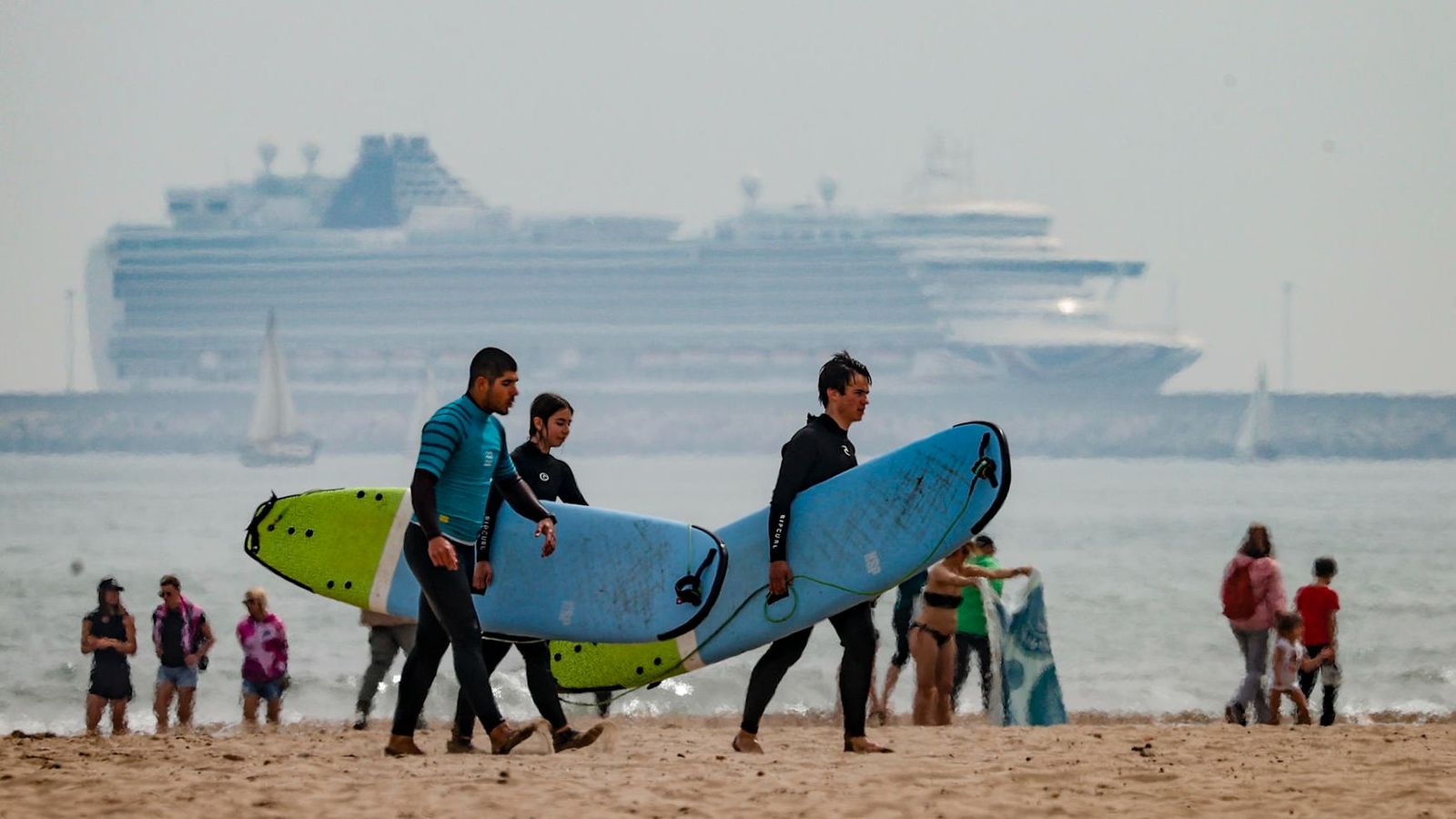 Tres joves surfistes ixen de la mar mentre un creuer de grans dimensions espera per a atracar al port de València