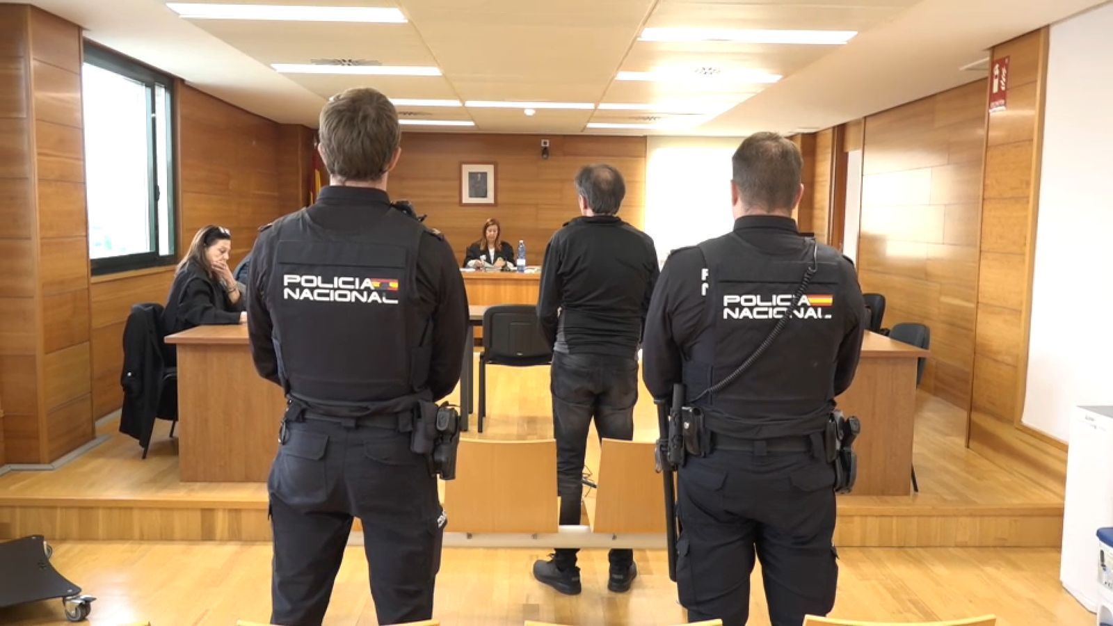 L'acusat, d'esquenes, custodiat per dos agents de Policia Nacional en el juí / À Punt NTC