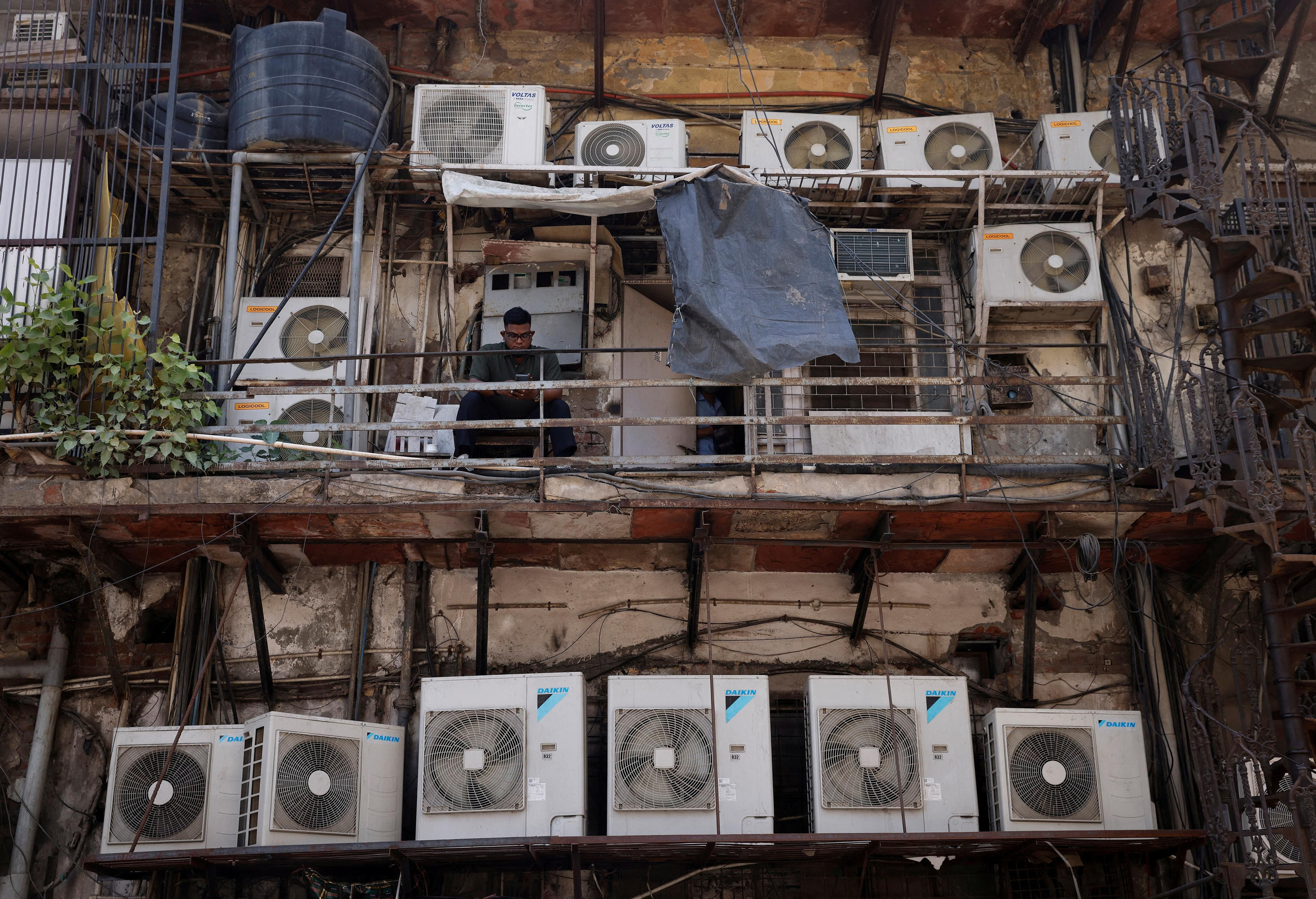 Un home assegut entre les unitats exteriors d'aire condicionat, en la part posterior d'un edifici comercial a Nova Delhi, l'Índia