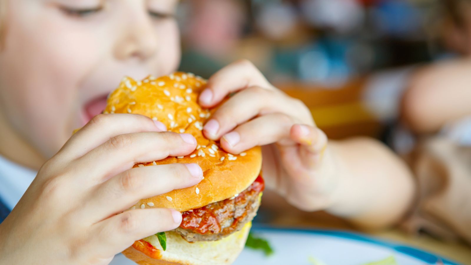 Un xiquet es menja una hamburguesa en una cadena d'alimentació / Shutterstock/ Irina Wilhauk
