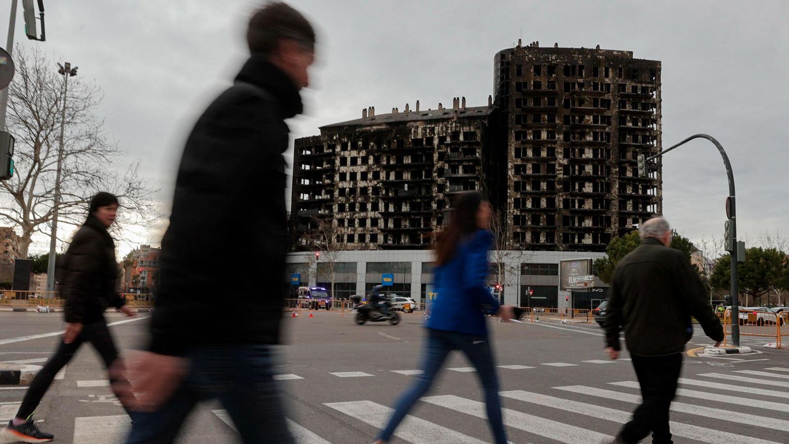 Diversos vianants creuen per un pas de peatons amb l'esquelet de l'edifici incendiat de fons, quatre dies després de l'incendi