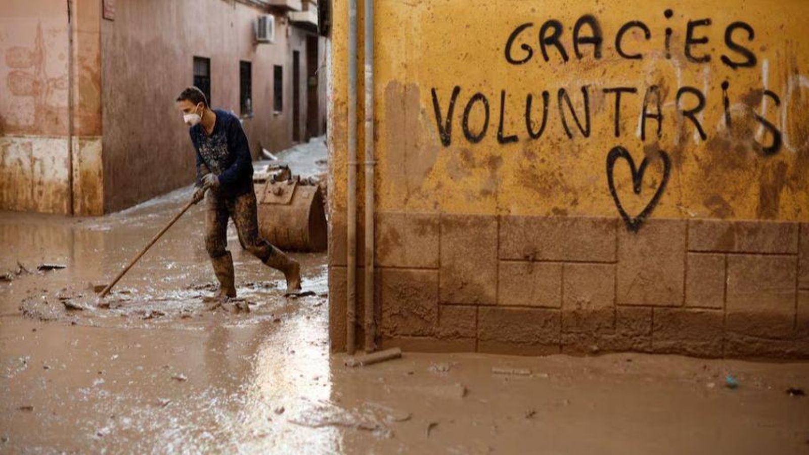 Un voluntari retira fang d'un carrer de Paiporta amb una pintada en la paret agraint el treball dels voluntaris